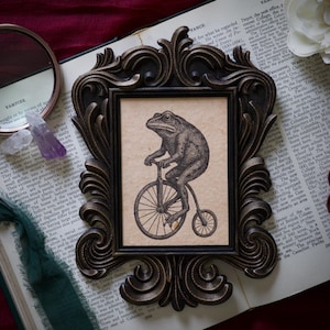 Może przedstawiać: Ozdobna, ciemnobrązowa rama z nadrukiem w stylu vintage przedstawiającym żabę na rowerze z dużym przednim kołem. Grafika umieszczona jest na beżowym tle, tworząc klasyczną, antyczną estetykę. Rama spoczywa na książce.
