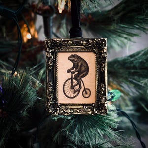 May include: A framed Christmas ornament featuring a vintage illustration of a frog riding a penny-farthing bicycle. The ornament has an ornate, dark gold frame and hangs from a black ribbon. The background is a blurred Christmas tree.