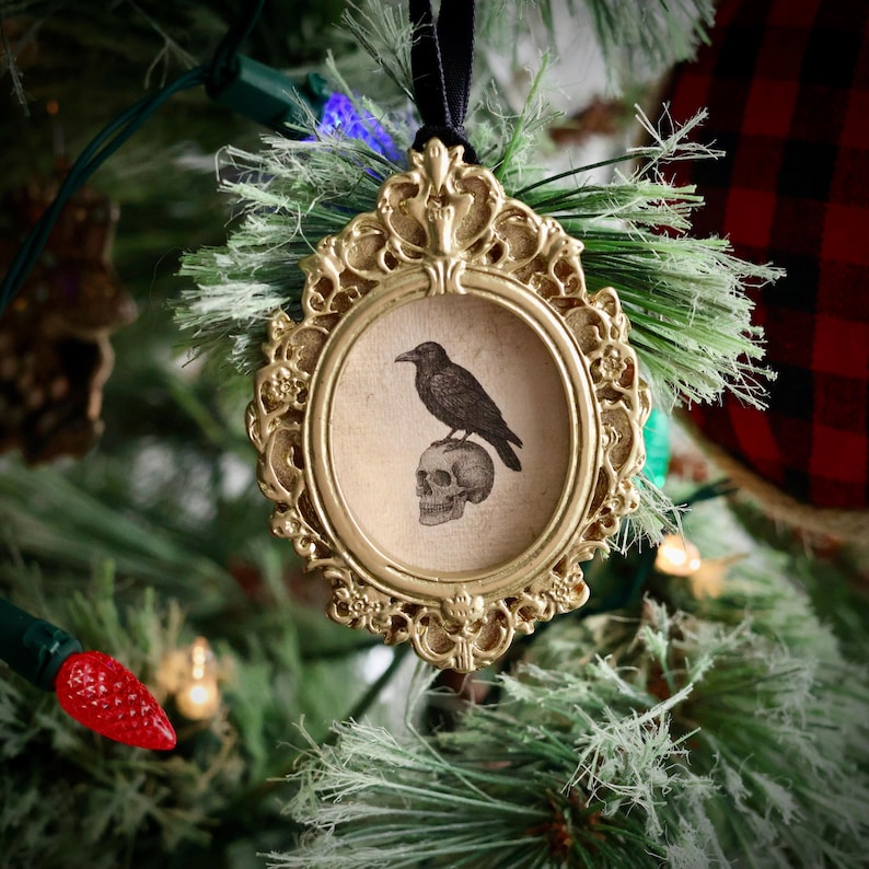 May include: A gold-colored, ornate framed ornament with a black raven perched on a skull. The ornament hangs from a black ribbon against a backdrop of a Christmas tree with green needles and red and white lights. The frame has an oval shape.