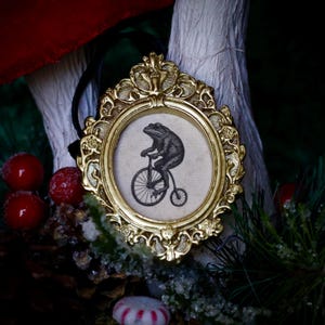 May include: A gold-colored, ornate framed ornament. The frame surrounds a circular image of a frog riding a penny-farthing bicycle. The frog is black and white. A black ribbon is attached to the top of the frame for hanging.