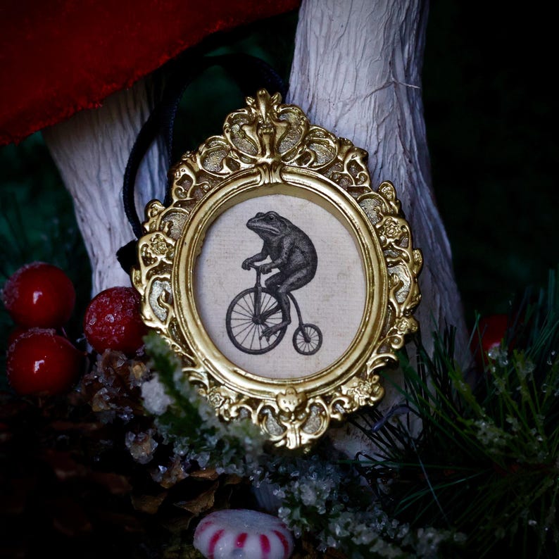 May include: A gold-colored, ornate framed ornament. The frame surrounds a circular image of a frog riding a penny-farthing bicycle. The frog is black and white. A black ribbon is attached to the top of the frame for hanging.