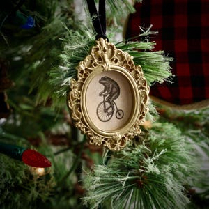 May include: A gold-colored, ornate framed Christmas ornament. The oval frame surrounds a sepia-toned image of a frog riding a penny-farthing bicycle. The ornament hangs from a black velvet ribbon and is displayed on a Christmas tree.
