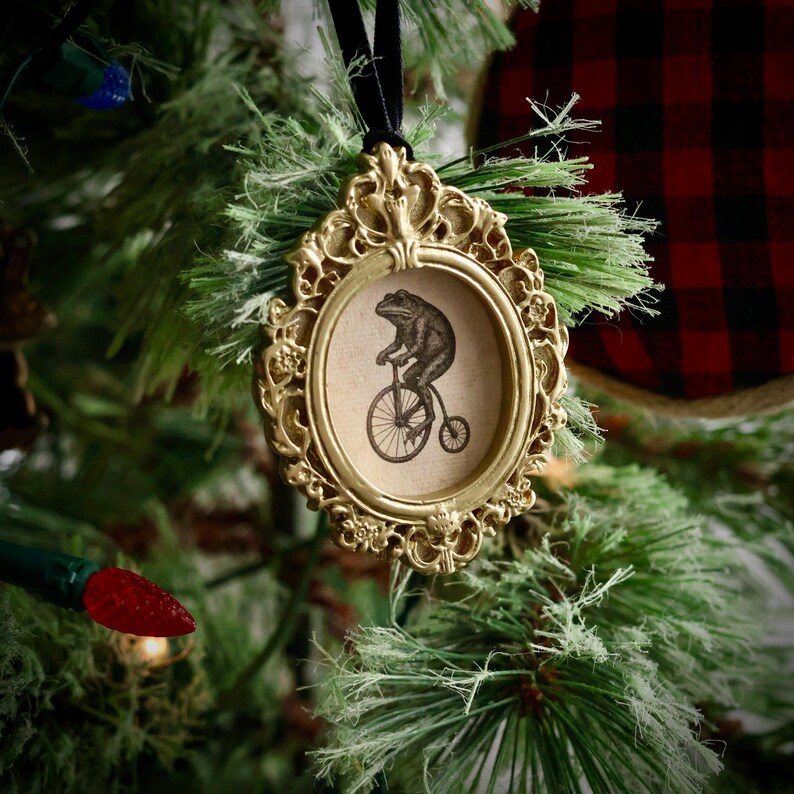 May include: A gold-colored, ornate framed Christmas ornament. The oval frame surrounds a sepia-toned image of a frog riding a penny-farthing bicycle. The ornament hangs from a black velvet ribbon and is displayed on a Christmas tree.