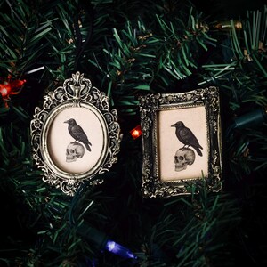 May include: Two ornate, antique-style picture frames with a raven perched on a skull image. One frame is oval, the other rectangular. Both are silver-toned and hang on a Christmas tree. The frames are likely ornaments with a gothic or Halloween theme.
