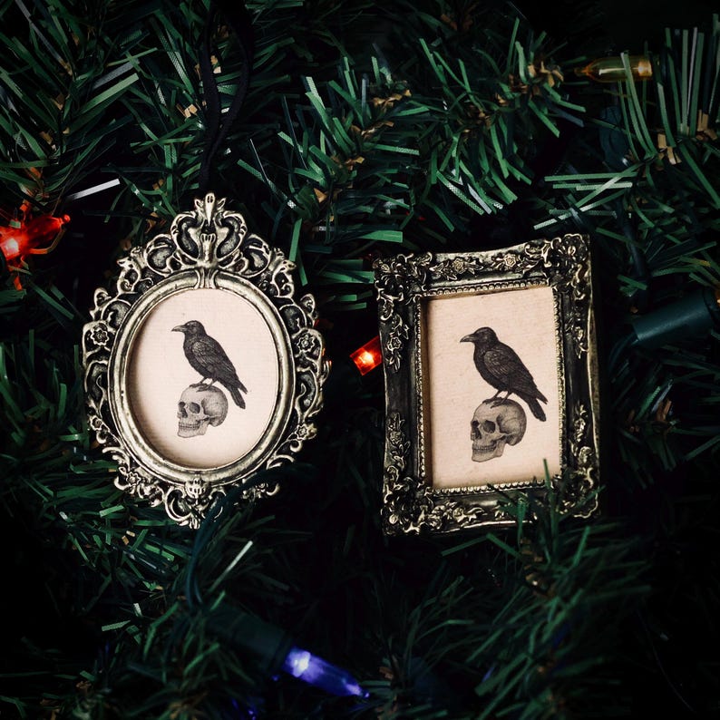 May include: Two ornate, antique-style picture frames with a raven perched on a skull image. One frame is oval, the other rectangular. Both are silver-toned and hang on a Christmas tree. The frames are likely ornaments with a gothic or Halloween theme.