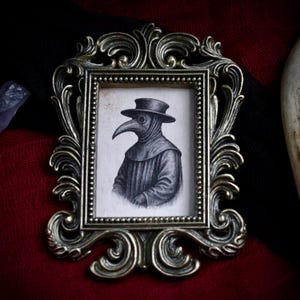 May include: Ornate, antique-style bronze frame with a small, detailed drawing of a plague doctor. The drawing is in black and white, depicting the doctor in traditional attire. The frame has intricate scrollwork and a beaded border.