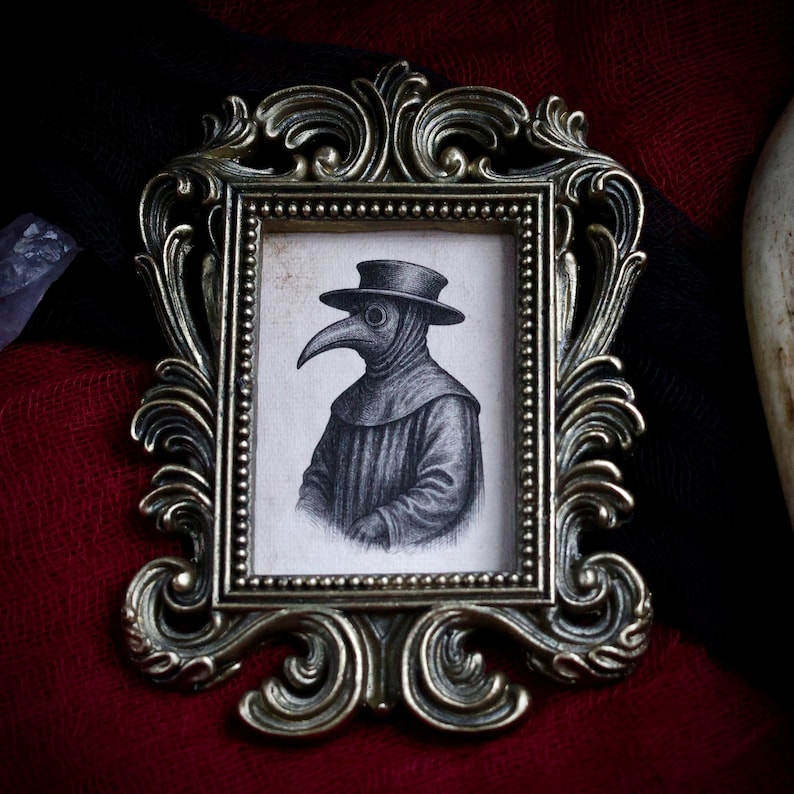 May include: Ornate, antique-style bronze frame with a small, detailed drawing of a plague doctor. The drawing is in black and white, depicting the doctor in traditional attire. The frame has intricate scrollwork and a beaded border.