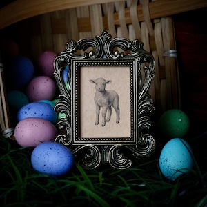 May include: An ornate, antique-style picture frame with a detailed lamb illustration. The frame is bronze-colored and rests on green grass, surrounded by colorful speckled Easter eggs in a basket. The image evokes a vintage, pastoral theme.