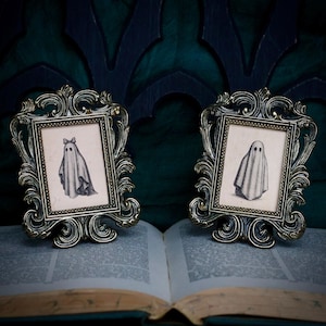 Peut inclure: Deux cadres dor&eacute;s anciens avec des d&eacute;tails orn&eacute;s. Chaque cadre contient une petite illustration en noir et blanc d'un fant&ocirc;me. Les cadres sont pos&eacute;s sur un livre ouvert aux pages vieillies, sur fond vert fonc&eacute;.