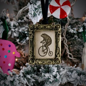 May include: A framed Christmas ornament with a vintage illustration of a frog riding a penny-farthing bicycle. The ornament has a gold-colored frame and a black velvet ribbon for hanging. The background is a snowy Christmas tree.