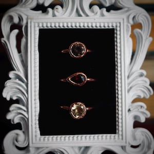 May include: Three gemstone rings displayed in a white ornate frame against a black background. The rings have copper-colored bands and feature a round, a teardrop, and a round-cut gemstone in shades of brown, red, and yellow.