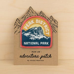 Könnte beinhalten: Ein aufbügelbarer, bestickter Aufnäher mit einer Berglandschaft und dem Text PEAK DISTRICT NATIONAL PARK. Der Aufnäher ist auf einer bergförmigen Pappunterlage befestigt, die mit dem Text IRON-ON adventure patch BY GOLDEN MOUNTAIN bedruckt ist.