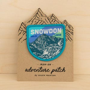 May include: A shield-shaped iron-on patch featuring a blue and white illustration of Snowdon, Wales, with the text "SNOWDON", "WALES", and "1,085M". The patch is mounted on a tan card with mountain line art and the text "IRON-ON", "adventure patch", and "BY GOLDEN MOUNTAIN".