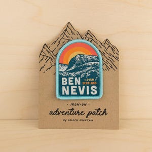 May include: An iron-on embroidered patch featuring a mountain landscape with a sunset, labelled Ben Nevis 1,345m Scotland. The patch is mounted on a brown cardstock backing shaped like mountain peaks with the text adventure patch by Golden Mountain.