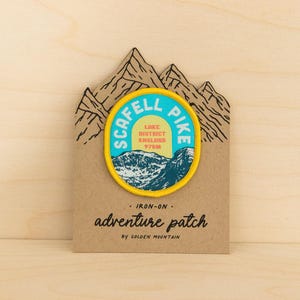 May include: A circular iron-on patch featuring a mountain landscape, the text SCAFELL PIKE, LAKE DISTRICT ENGLAND 978M, and a yellow border. The patch is mounted on a brown card shaped like mountain peaks with the text adventure patch.