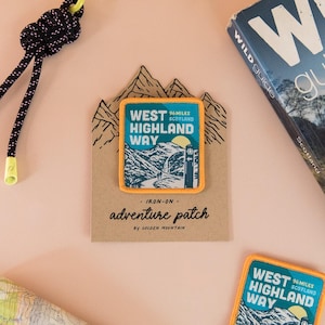 May include: Two rectangular iron-on patches featuring a West Highland Way graphic with mountains, a trail marker, and a yellow border. One patch is attached to a mountain-shaped cardboard backing with the text Iron-on adventure patch.
