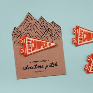 May include: Embroidered orange and white "Happy Camper" pennant patches. One patch is attached to a brown card with a mountain illustration and the text "adventure patch". The patches are on a light blue background.