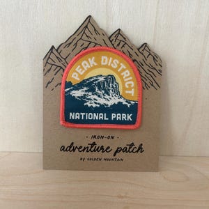 May include: An iron-on embroidered patch featuring a mountain landscape with the text PEAK DISTRICT NATIONAL PARK. The patch has a coral-colored border and is mounted on a brown card shaped like mountain peaks with the text adventure patch.