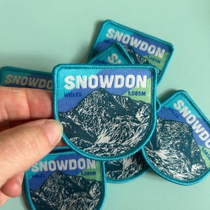 May include: Embroidered patches featuring a mountain landscape with the text "SNOWDON WALES 1,085M" in white and teal. The patches have a teal border and a blue and teal mountain design.