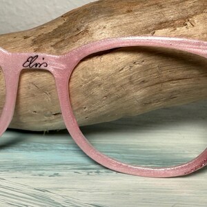 May include: Pink eyeglasses with a glittery finish and the name "Elvis" written on the frame. The right temple features a small, stylized face. The glasses are set against a wooden background.
