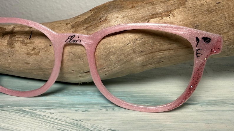 May include: Pink eyeglasses with a glittery finish and the name "Elvis" written on the frame. The right temple features a small, stylized face. The glasses are set against a wooden background.