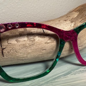 May include: Cat-eye spectacles with a colour gradient from green to red to magenta. The left side of the frame is decorated with a rainbow gem and clear rhinestones. The glasses are displayed on a piece of driftwood.