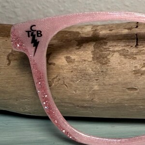 May include: Close-up of a pink eyeglass frame with glitter accents. The frame has the letters "C T4 B" and a lightning bolt symbol in black. The frame is curved and appears to be made of a translucent material.
