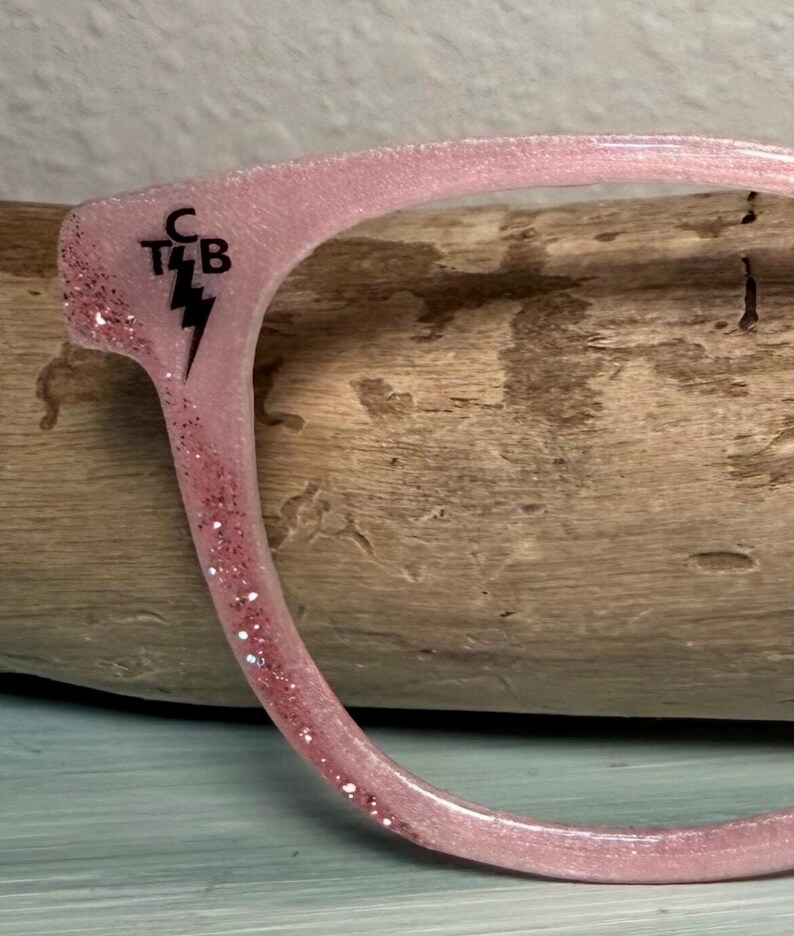 May include: Close-up of a pink eyeglass frame with glitter accents. The frame has the letters "C T4 B" and a lightning bolt symbol in black. The frame is curved and appears to be made of a translucent material.