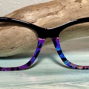 May include: Cat-eye style eyeglasses with a black frame and purple accents. The temples feature a purple and black pattern, and the corners are adorned with sparkly purple floral embellishments. A stylish accessory.