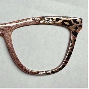 May include: Close-up of a pair of eyeglasses with a glittery rose gold frame. The upper right corner features a leopard print design with rhinestones. The frame is embellished with small, clear rhinestones.