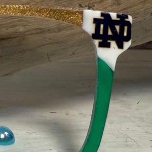 Könnte beinhalten: Ein dekorativer Haken mit einem grünen, gebogenen Körper. Die Oberseite hat einen weißen Abschnitt mit den marineblauen Buchstaben "ND" und einen goldenen Glitzerakzent. Der Haken dient wahrscheinlich zum Aufhängen von Gegenständen.