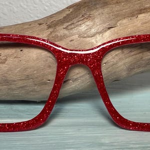 May include: Red glitter eyeglasses frames with a rectangular shape. The frames are a vibrant red color and are covered in sparkling glitter. The glasses are set against a wooden background.