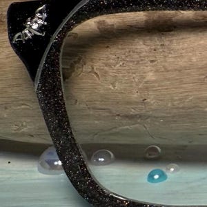 Puede incluir: Primer plano de una montura de gafas negra con purpurina y un emblema plateado. La montura está sobre un fondo de madera. La lente es transparente y hay varios objetos redondos y coloridos en primer plano.