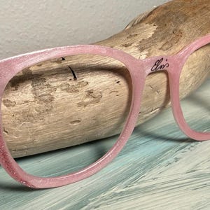 May include: Pink eyeglasses with glitter accents and the word "Elvis" on the frame. The left arm of the glasses has the letters "C T&B" with a lightning bolt. The glasses are resting on a piece of driftwood.