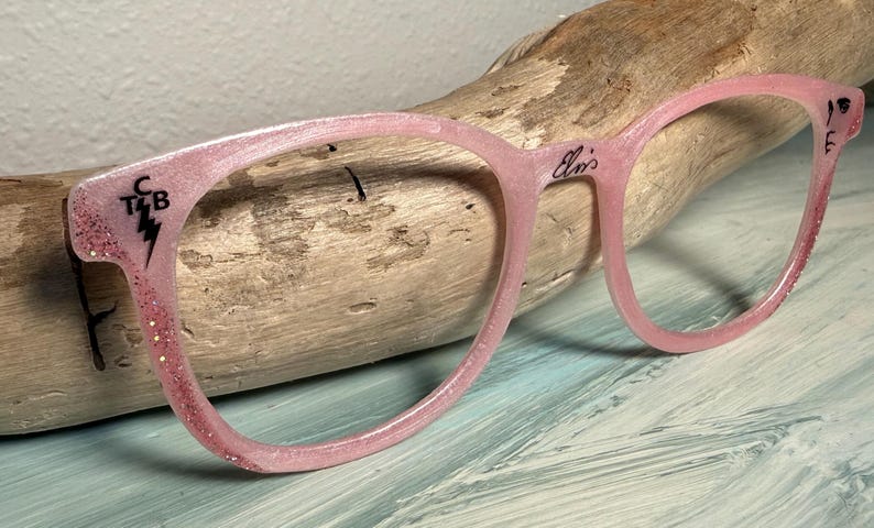 May include: Pink eyeglasses with glitter accents and the word "Elvis" on the frame. The left arm of the glasses has the letters "C T&B" with a lightning bolt. The glasses are resting on a piece of driftwood.