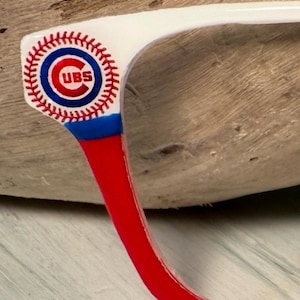 Puede incluir: Primer plano de una montura de gafas blanca con un diseño de béisbol rojo y azul. La montura presenta el logotipo de los Chicago Cubs. La patilla de las gafas es roja, pasando a azul y blanco en la bisagra.