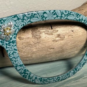 May include: Teal-colored eyeglasses with a floral lace pattern. The frames feature a decorative flower with pearl and gold accents on the upper left corner. The glasses are set against a wooden background.