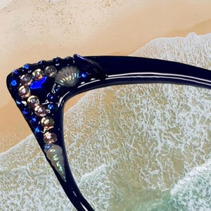 May include: Dark blue eyeglasses with decorative embellishments. The frame is adorned with iridescent gems, seashells, and blue accents. The glasses are set against a backdrop of a sandy beach and ocean waves.