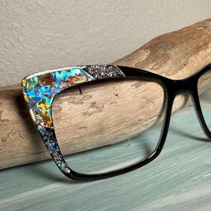 Puede incluir: Gafas con montura negra con un diseño único en la parte superior de la montura. El diseño presenta piezas de vidrio iridiscentes y coloridas y detalles de purpurina plateada. Las gafas descansan sobre un trozo de madera flotante.