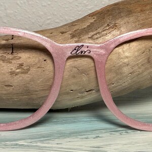 May include: Pink eyeglasses with a glittery finish. The frames have a round shape and feature the name "Elvis" on the bridge. The left side has the initials "T&B" with a lightning bolt, and the right side has a small face illustration.