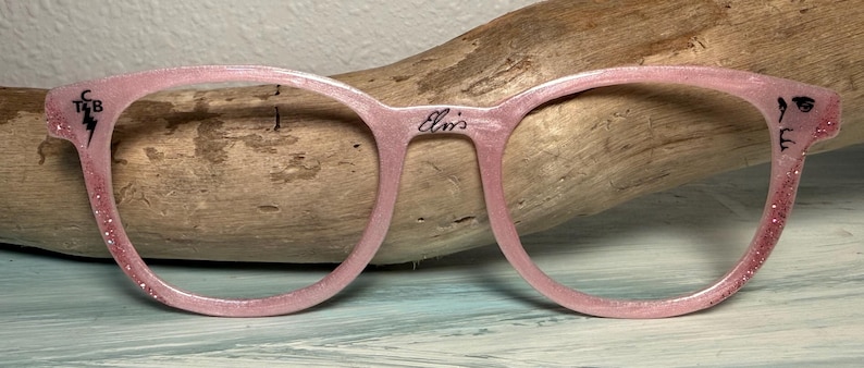 May include: Pink eyeglasses with a glittery finish. The frames have a round shape and feature the name "Elvis" on the bridge. The left side has the initials "T&B" with a lightning bolt, and the right side has a small face illustration.