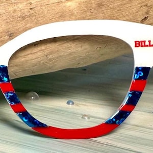 May include: A white eyeglass frame with red and blue accents. The frame has the word "BILLS" in red letters. The frame is decorated with red stripes and blue patterns. The frame is on a light blue surface.