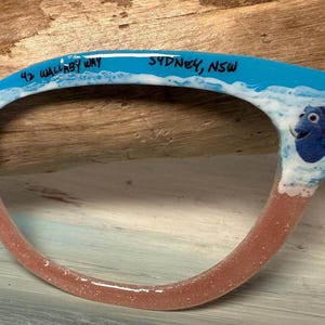 May include: A pair of eyeglasses with a blue frame, featuring the text "42 Wallaby Way Sydney, NSW". The frame has a wave design with a Dory character from Finding Nemo. The lower frame is a glittery pink.