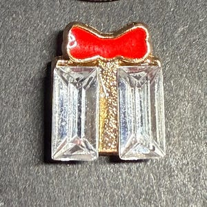 May include: A gold-toned gift box charm with a red bow on top. The gift box has two rectangular, clear crystal-like stones. The charm is likely for a bracelet or necklace.