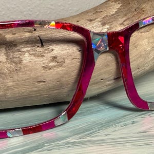 May include: A pair of eyeglasses with a vibrant, colorful design. The frames are a mix of red and pink, with iridescent accents. The glasses are set against a natural wood background.