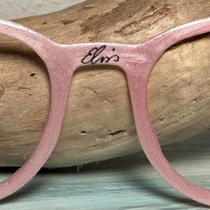 May include: Close-up of pink eyeglasses with the text "Elvis" in cursive script on the bridge. The frames have a subtle glitter effect. The glasses are set against a wooden background.