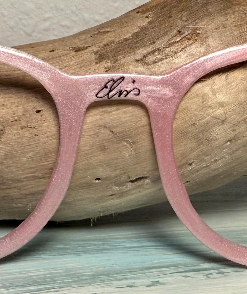 May include: Close-up of pink eyeglasses with the text "Elvis" in cursive script on the bridge. The frames have a subtle glitter effect. The glasses are set against a wooden background.