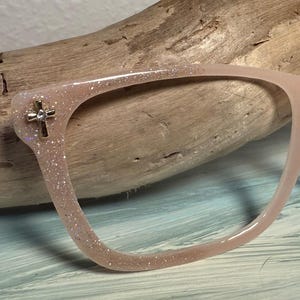 May include: A pair of light pink eyeglasses with a glittery finish. The frames feature a small silver cross embellishment on the outer corner. The glasses are set against a wooden background.