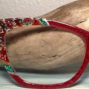 Pode incluir: Armação de óculos vermelha com glitter com detalhes decorativos dourados, prateados e verdes. A armação tem formato de olho de gato. Os óculos estão em um fundo de madeira.
