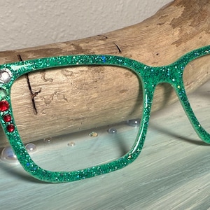 Op de afbeelding: Groene glitterbril met decoratieve rode en heldere strass-steentjes aan de linkerkant. De bril staat tegen een houten achtergrond en een lichtblauwe ondergrond, waardoor een levendig en opvallend accessoire ontstaat.